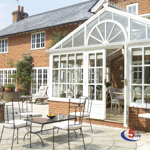 A bright and inviting conservatory attached to a brick house, featuring large glass windows and doors, surrounded by a stone patio with outdoor seating.